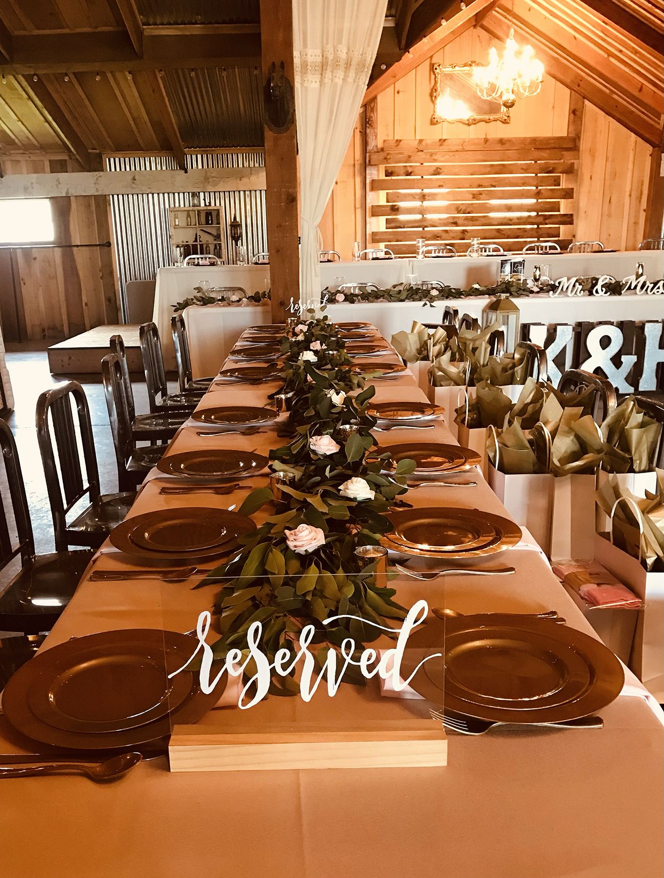 Long reception table in the barn with gold chargers, a greenery and rose runner and a reserved sign, with the head table behind
