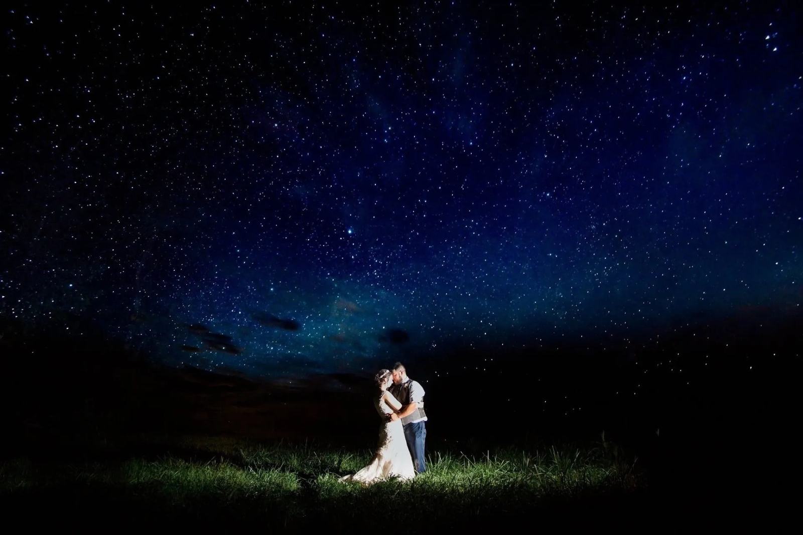 Bride and groom embracing in a field at night under a sky full of stars