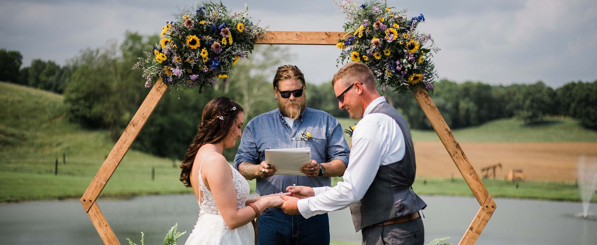 Bride and groom exchanging rings under a wooden hexagon arch topped with sunflowers beside the pond